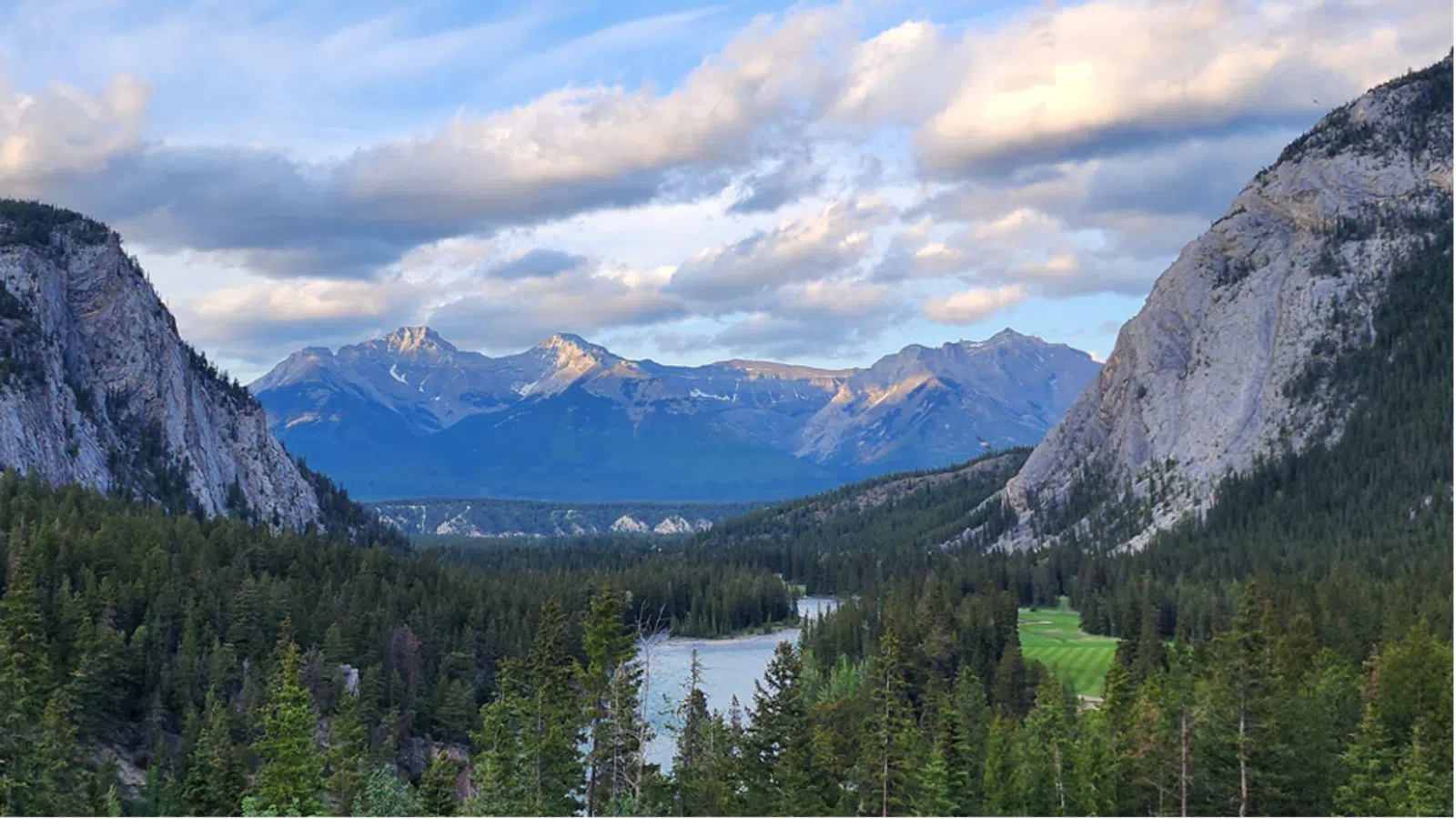 River winding through dense forest with towering mountains and cloudy blue sky in the background