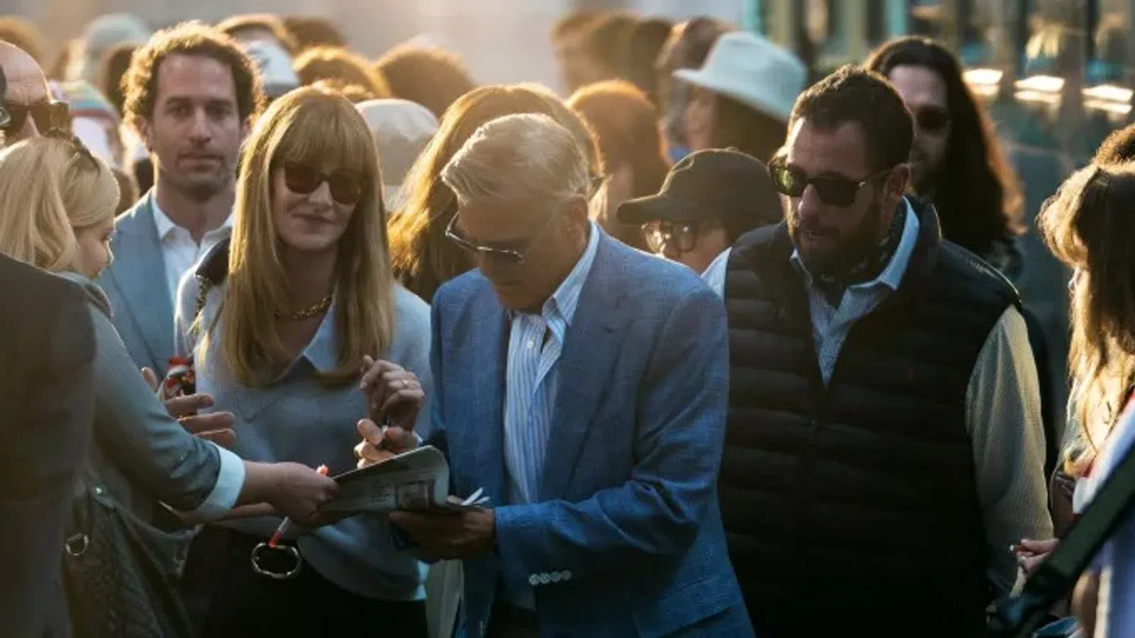 Celebrity signing autographs for fans outdoors in stylish attire during daytime event with crowd around.