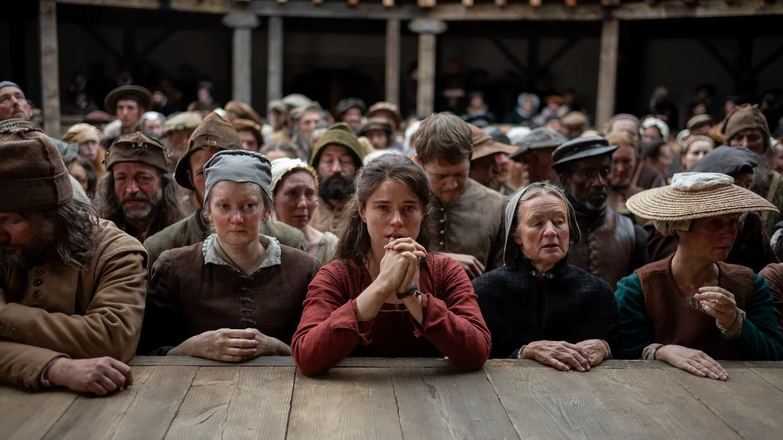 Crowd of somber medieval villagers in simple clothing gathered around a wooden table in a rustic hall.