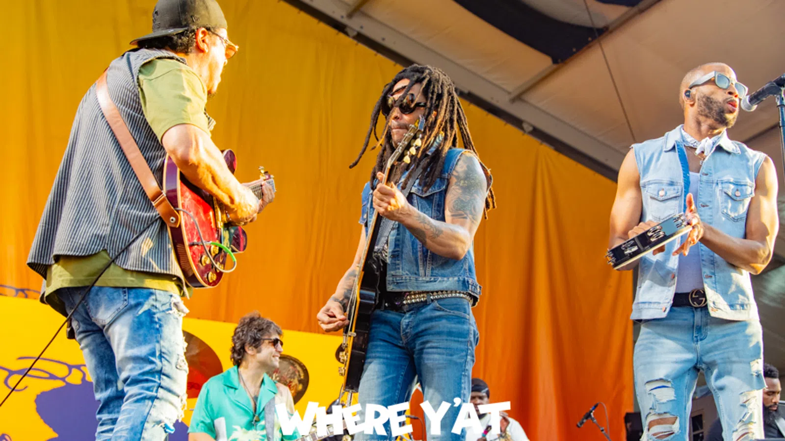 Musicians perform energetically on stage with guitars, tambourine, and drums in front of a yellow backdrop.