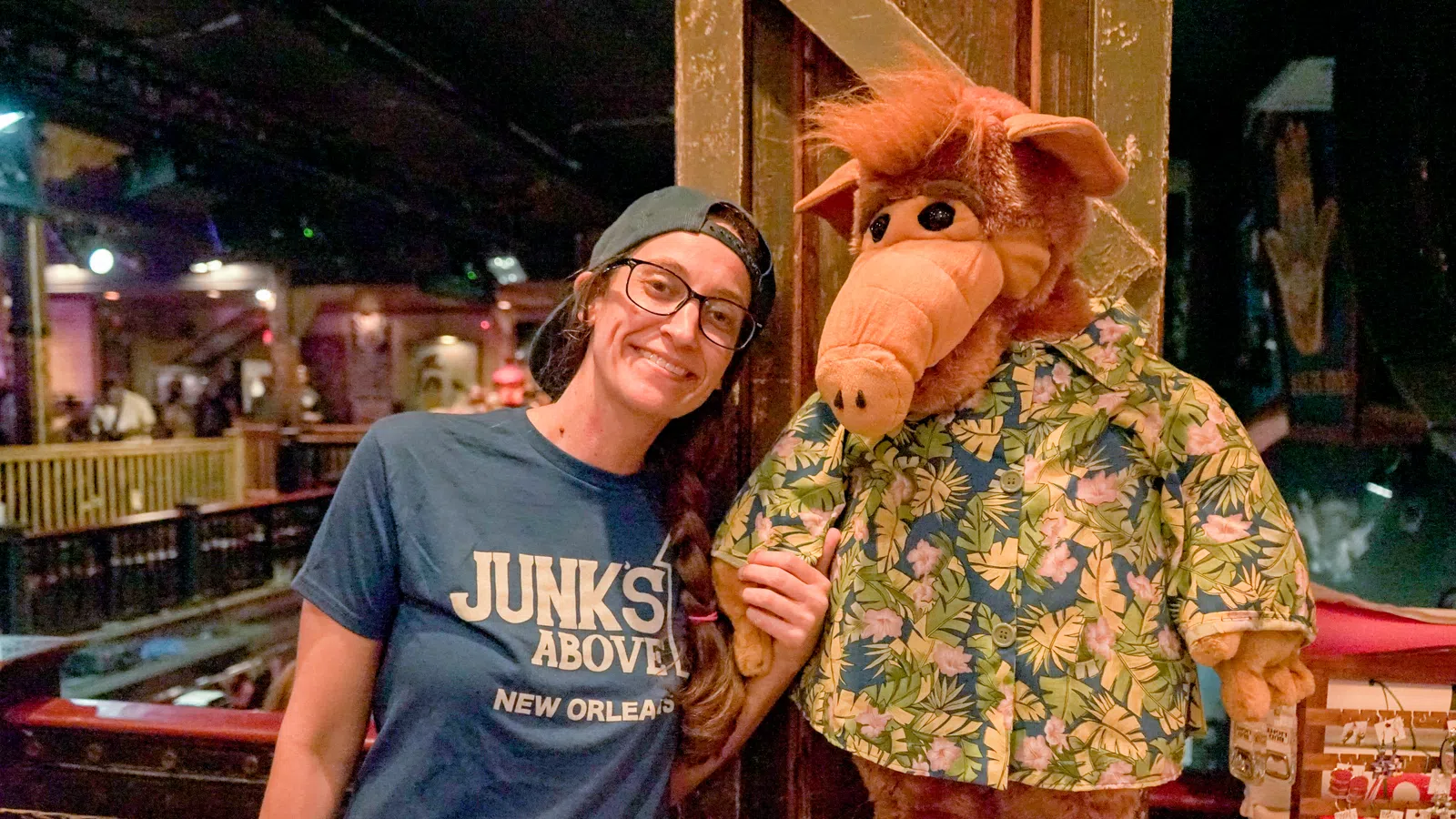 Woman in glasses and cap posing with ALF character wearing a tropical shirt inside a cozy venue.