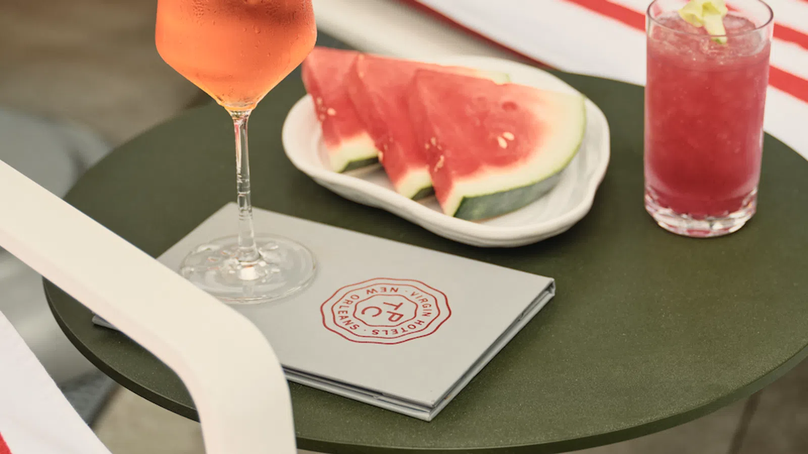 Glass of rose wine, plate with watermelon slices, pink cocktail, and menu on outdoor green table.