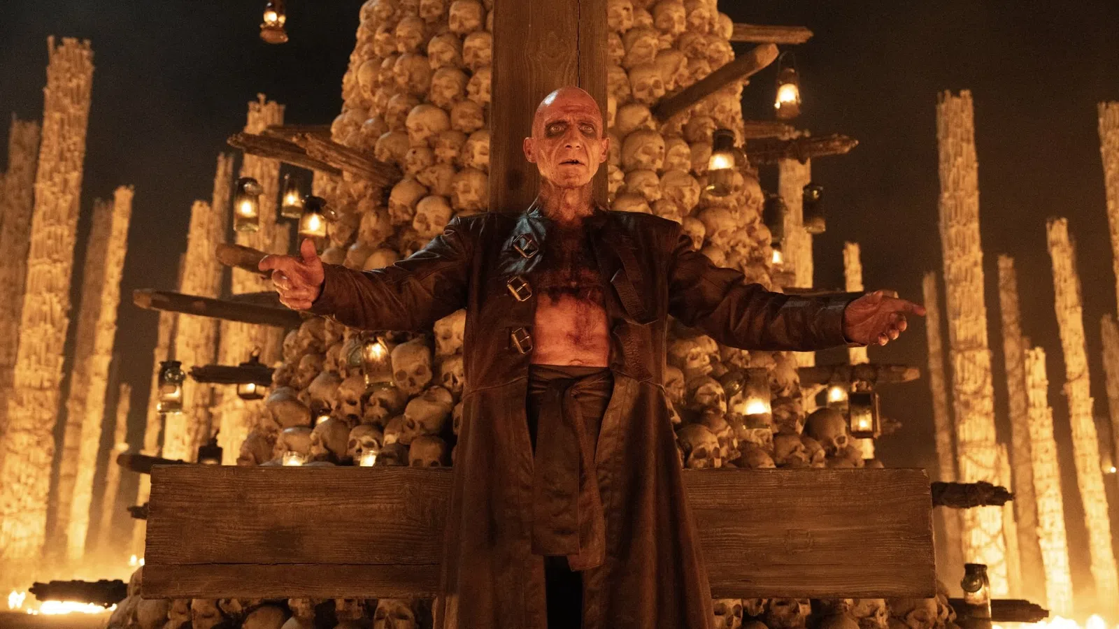 A bald man with scars stands in front of a towering pile of skulls and lit lanterns, spreading his arms wide.