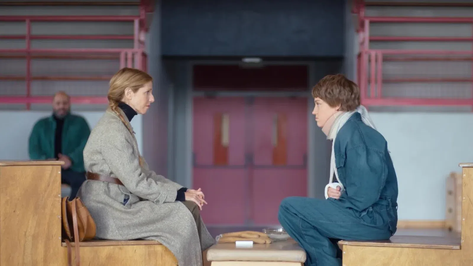 Two women seated on wooden benches facing each other in a stark room, engaged in serious conversation.