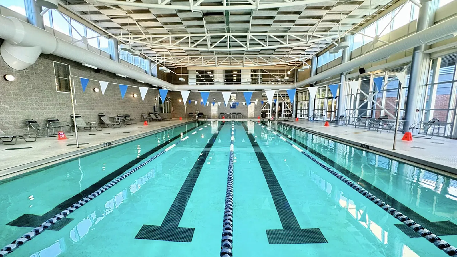 Indoor swimming pool with clear lanes, flags, deck chairs, and large windows letting in natural light.