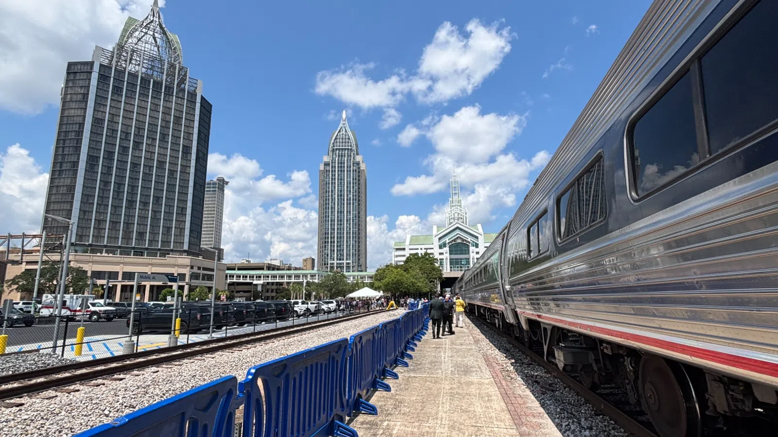 Silver train at platform with blue seats and city skyscrapers under a partly cloudy sky.