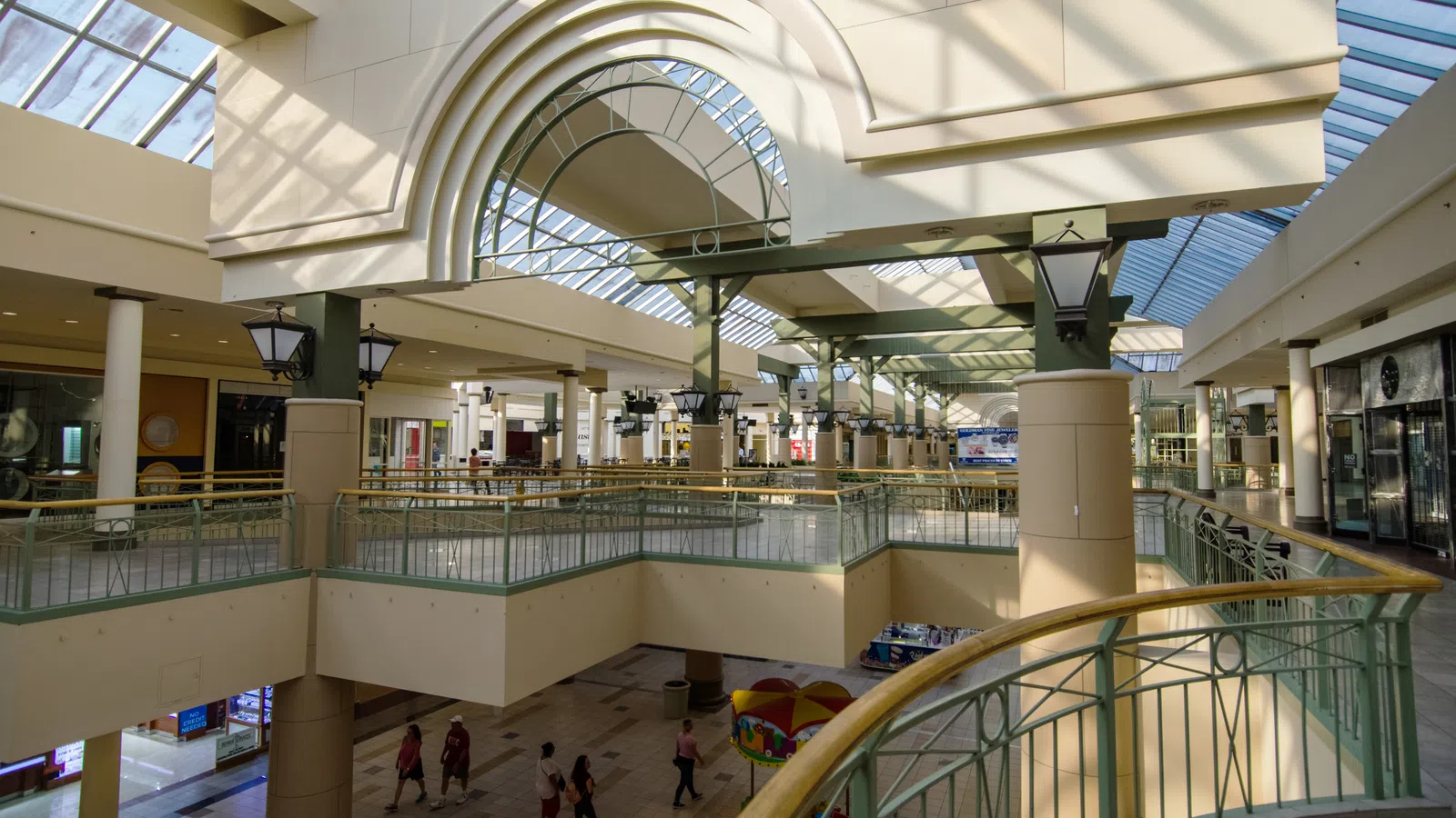 Spacious bright shopping mall interior with glass ceiling, columns, railings, and people walking below