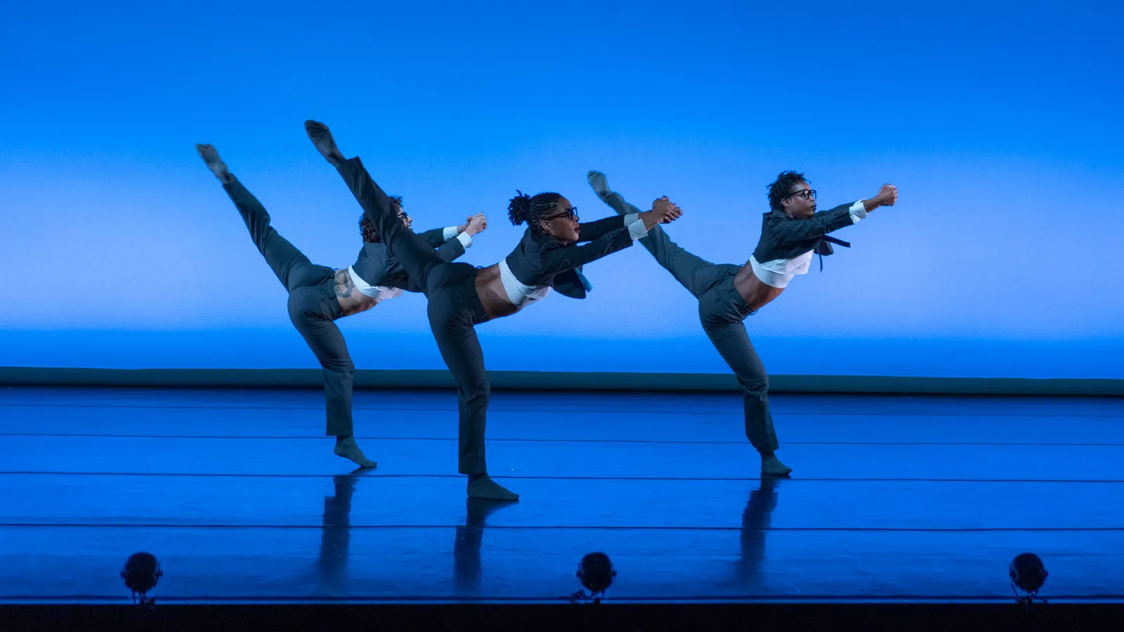 Three dancers in gray suits perform a high leg balance on a blue-lit stage in unison.
