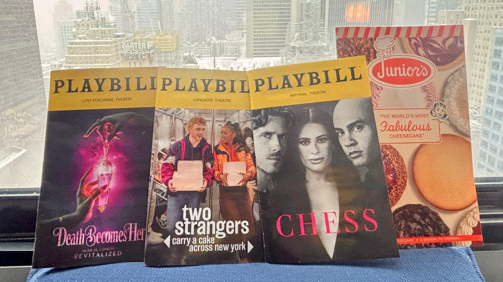 Three Playbill theatre programs and a Junior's cheesecake menu displayed on a windowsill with a snowy city skyline in the background.