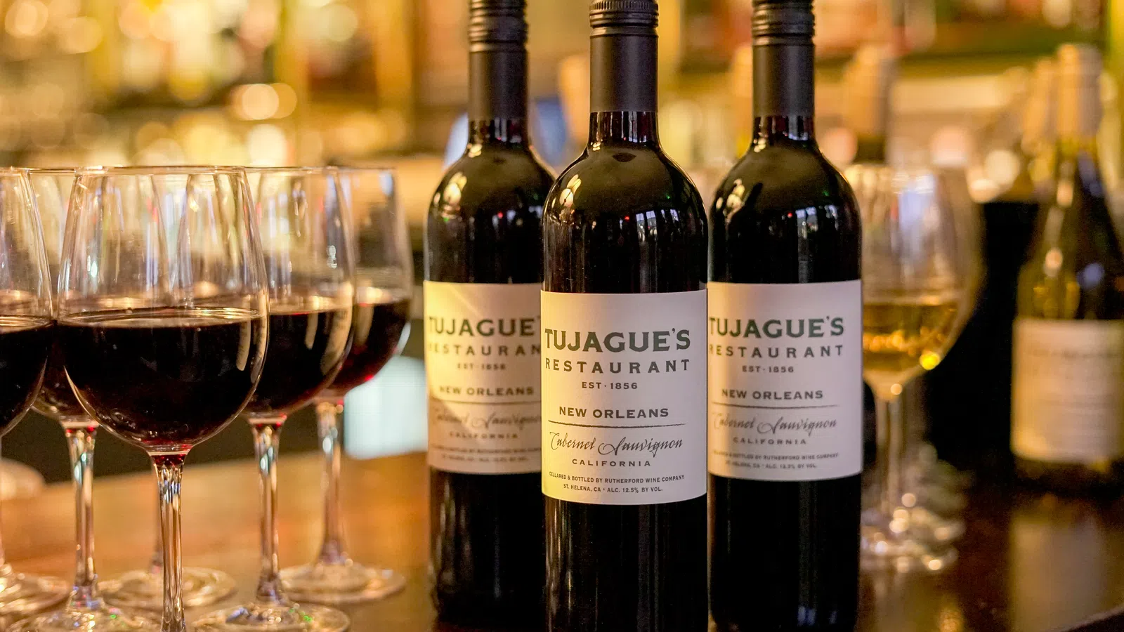 Three bottles of Tujague's Restaurant red wine with six filled wine glasses on a wooden bar counter.