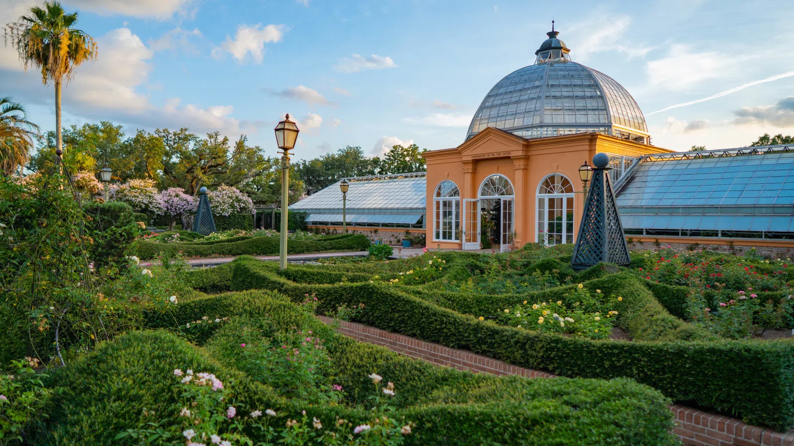 Historic glass-domed conservatory with manicured garden and hedges under a blue sky with clouds.