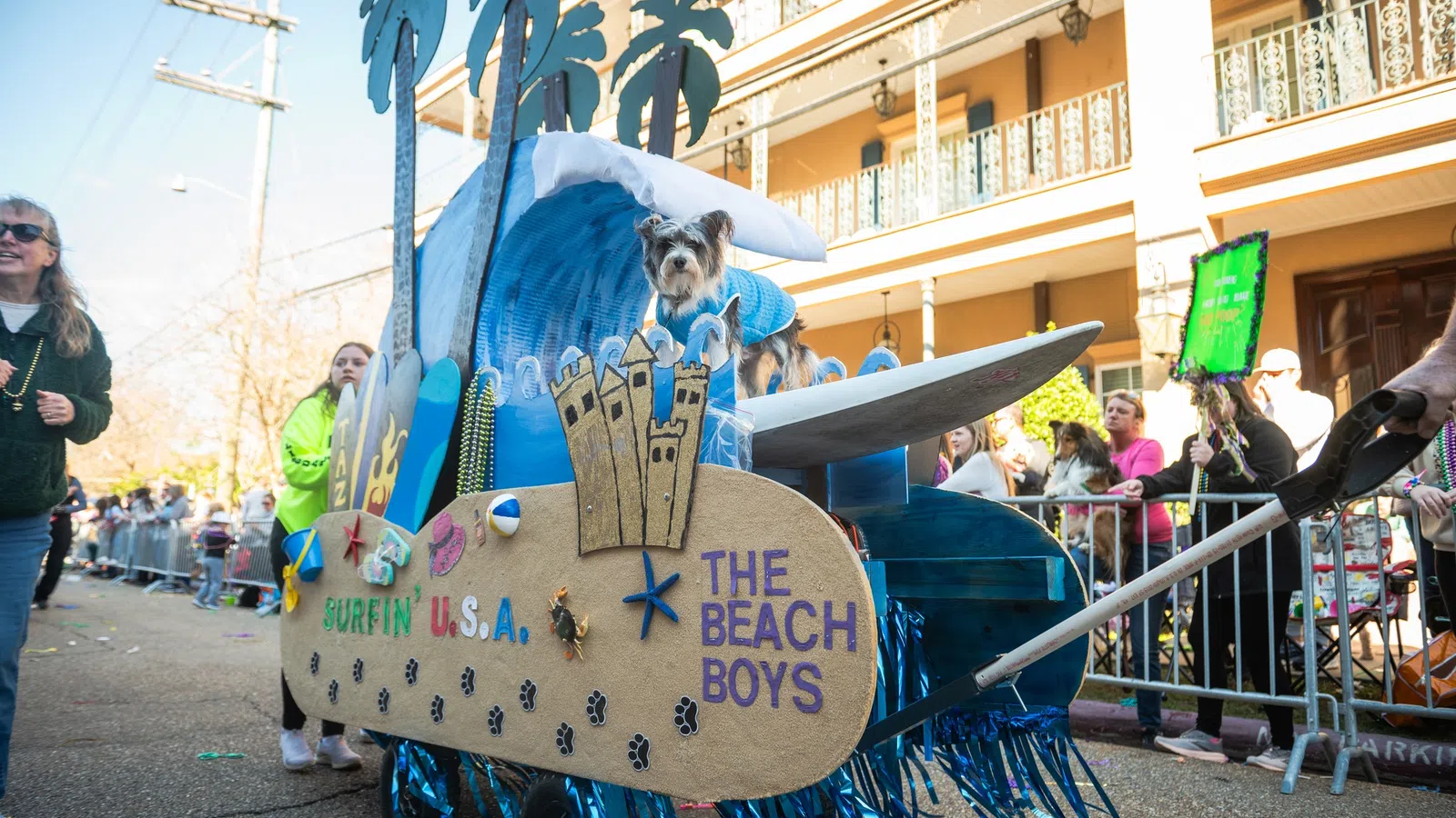Dog in blue sweater on a beach-themed parade float with surfboard and palm tree decorations during street event
