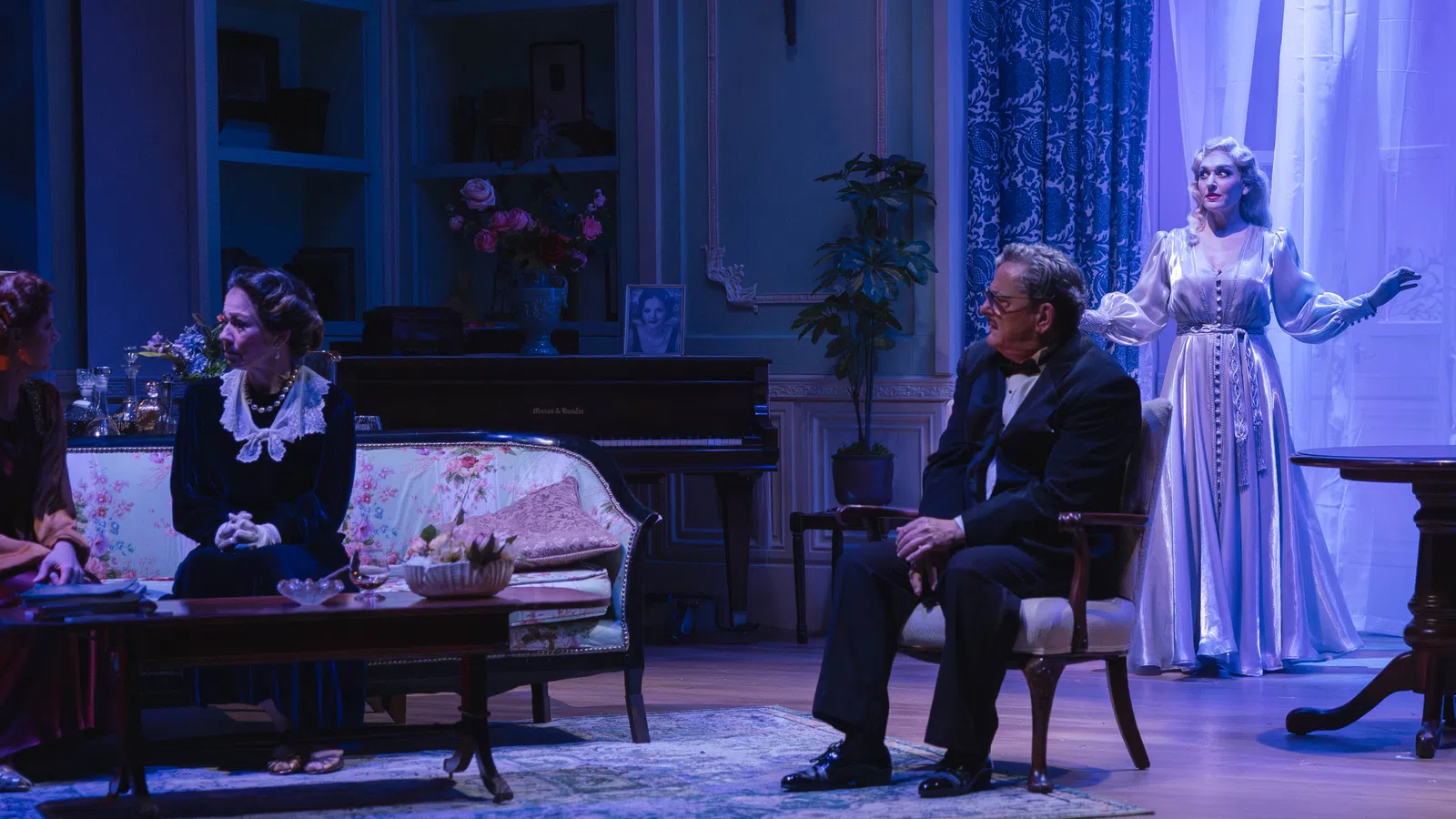 Theater scene with four actors in vintage attire on an elegant living room stage set with blue lighting.