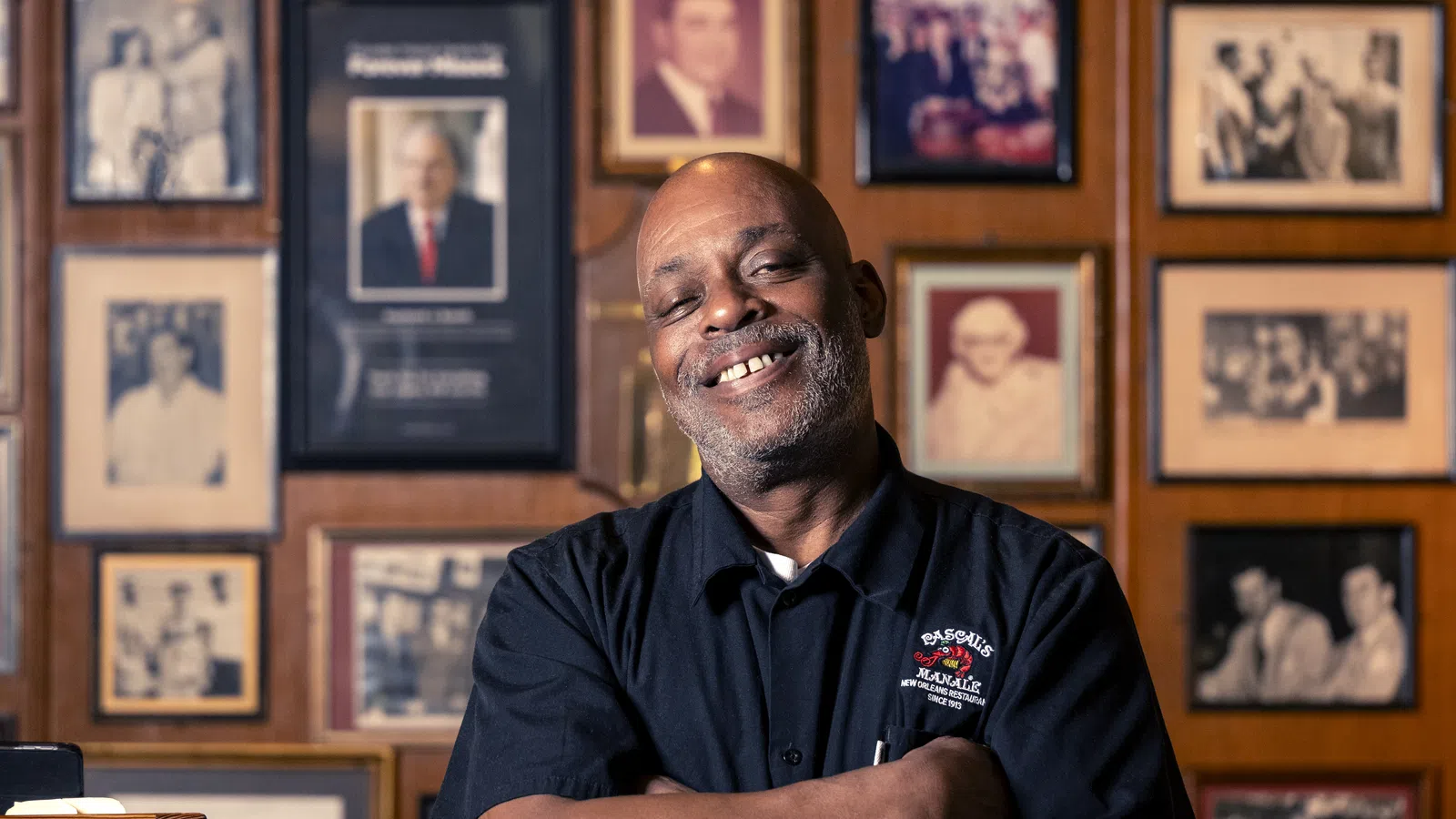 Smiling man with folded arms standing in front of a wall covered with framed vintage photographs.