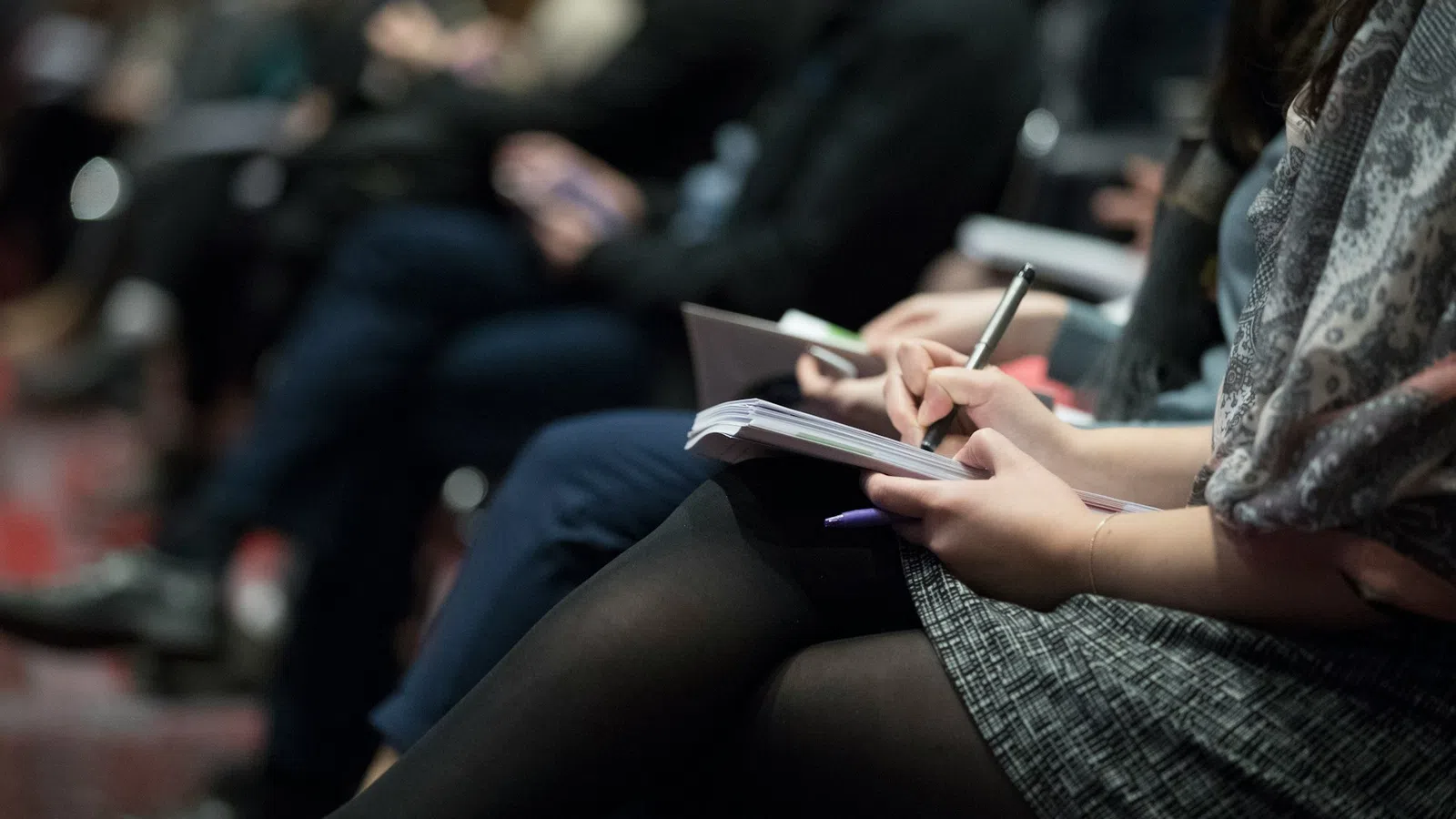Participants engaged in a conference, taking notes in notebooks during a presentation.