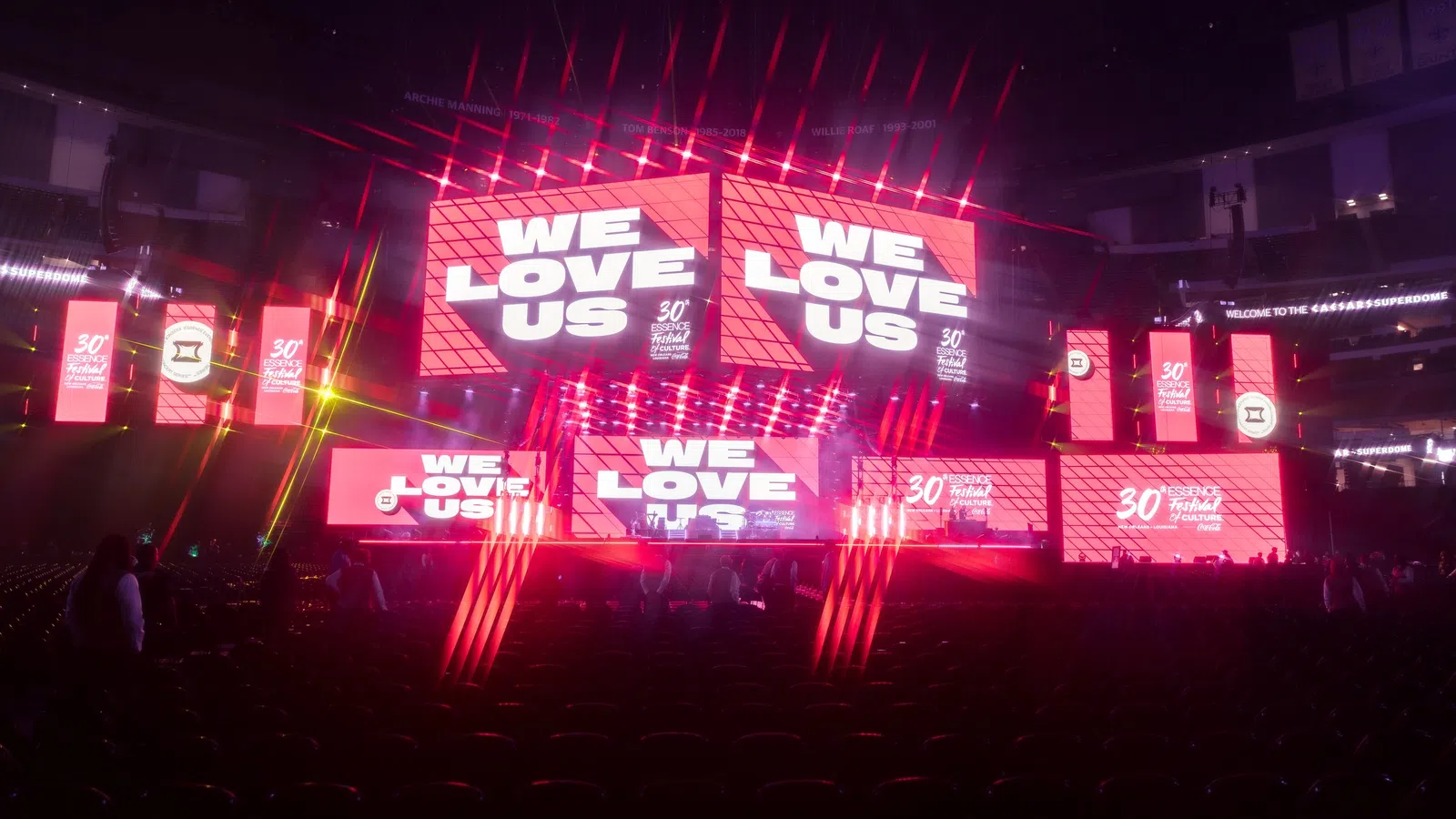 Vibrant stage setup with neon lights and large screens displaying 'WE LOVE US' at a lively event.