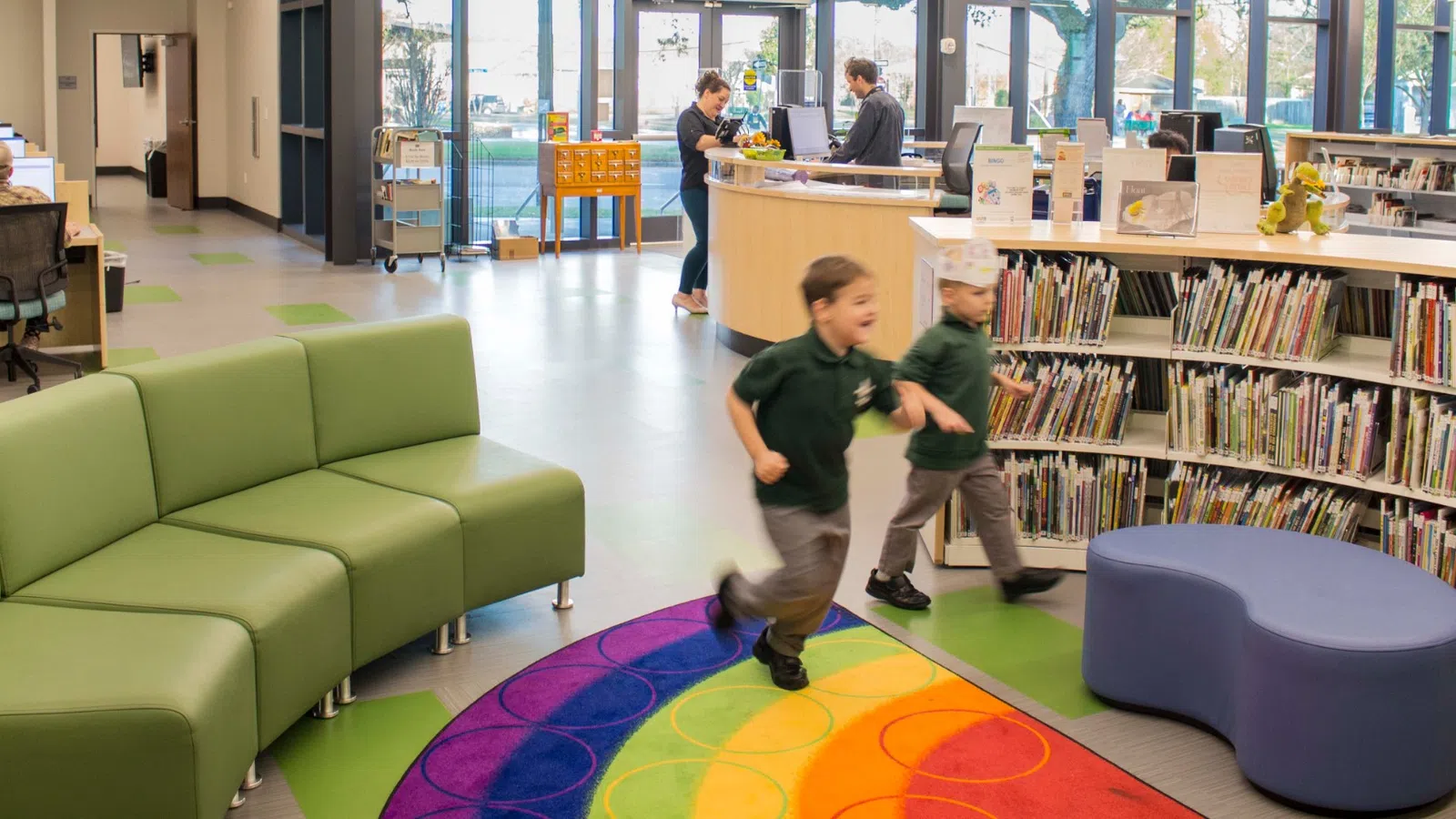 Children running on a colorful carpet in a modern library with bookshelves, seating, and large windows in the background