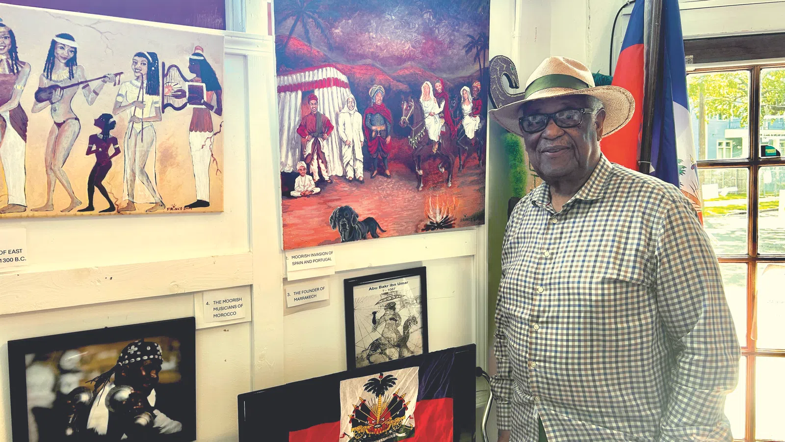 Elderly man in a straw hat stands next to Haitian flag and cultural paintings inside a bright gallery space.