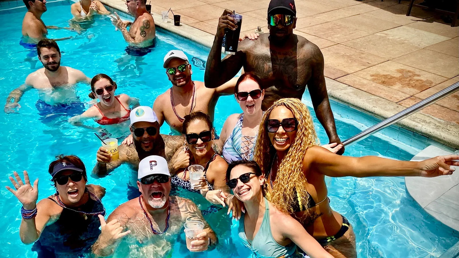 Group of happy adults enjoying drinks and fun in a sunny outdoor swimming pool party.