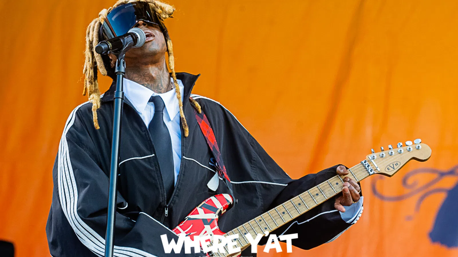 Musician performing on stage, wearing a black jacket and playing a vibrant guitar.