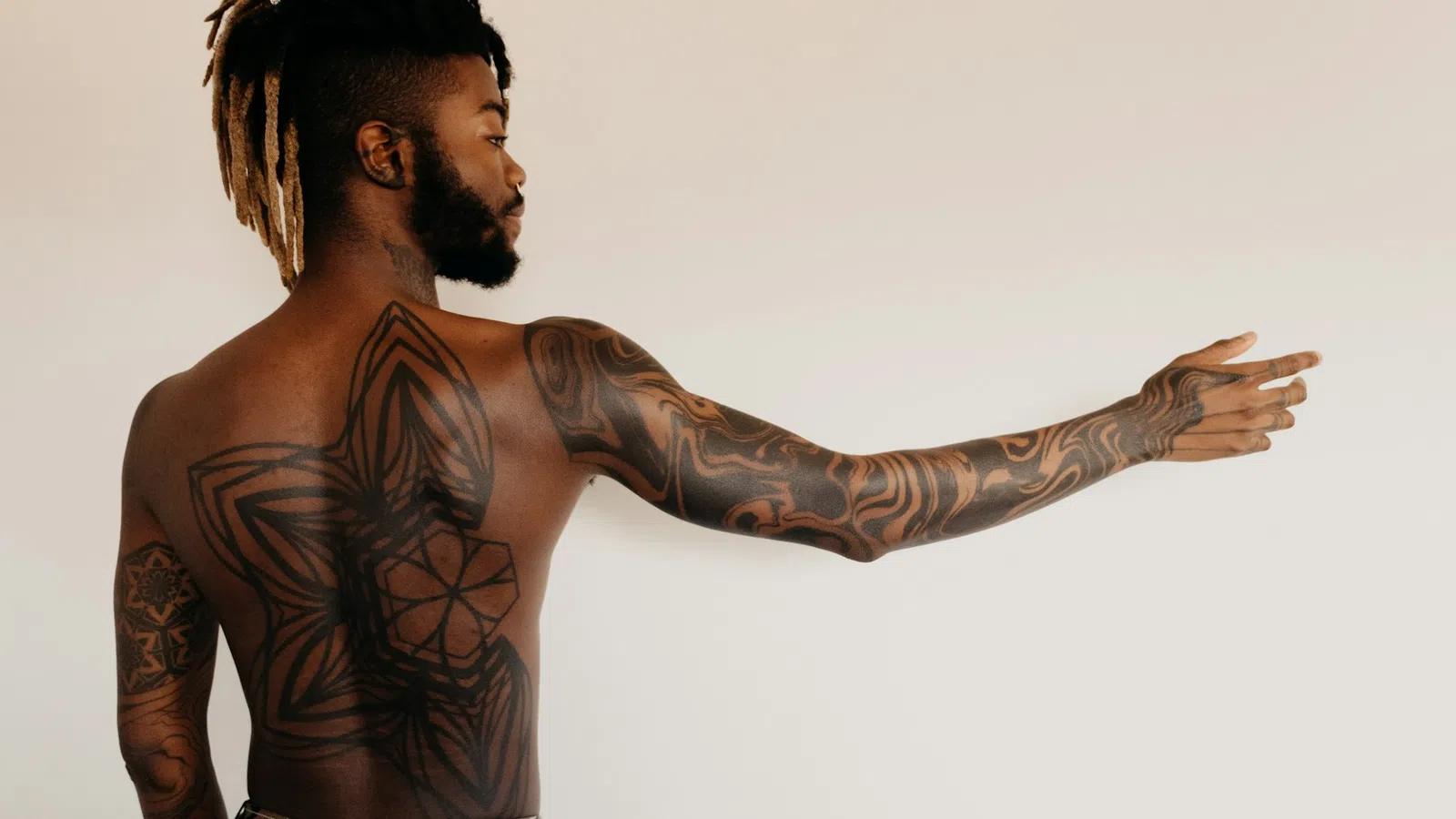 Man with detailed geometric tattoos covering his back and arm, pointing to the side against a plain background.