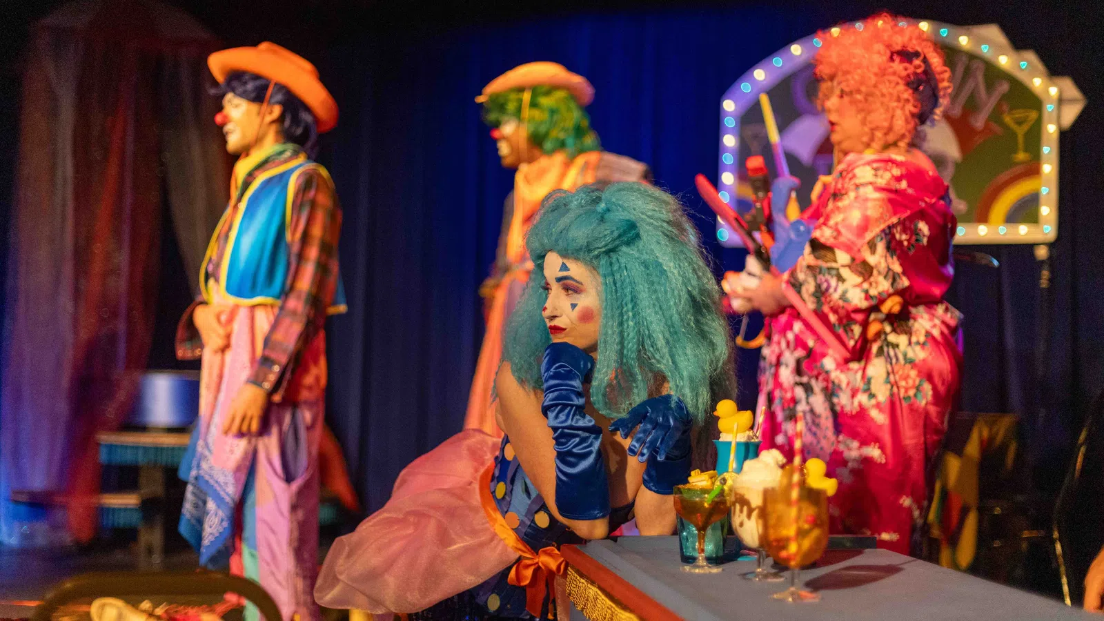 Colorful clowns in vibrant costumes perform on stage with drinks on a table under theatrical lighting.