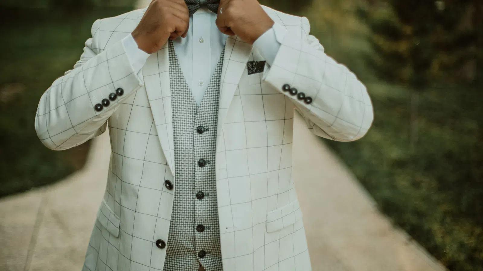 Man adjusting bow tie wearing a white checkered suit jacket and houndstooth vest outdoors on pathway.