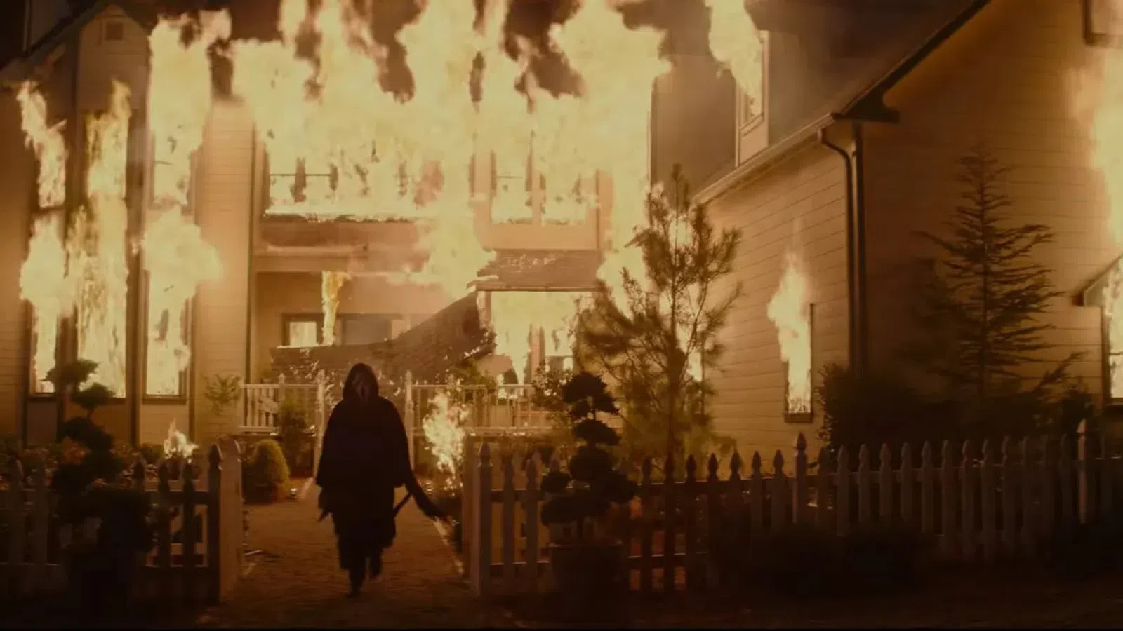 Person walks towards a large house engulfed in flames at night, amidst a white picket fence and trees.