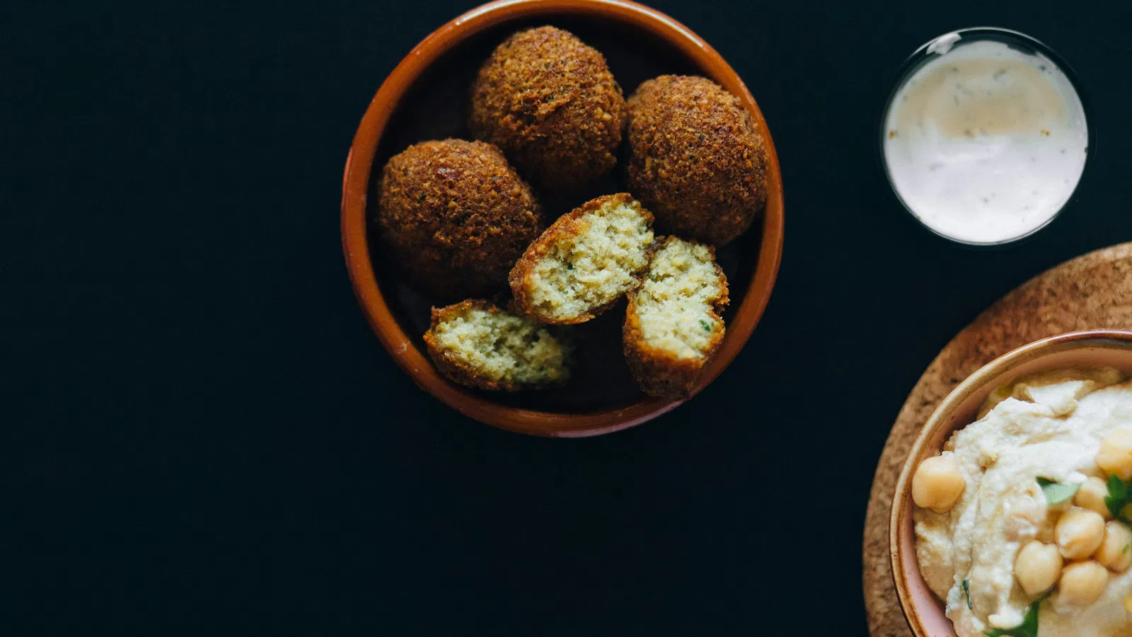Bowl of golden falafel balls with cut pieces showing inside, served with creamy dip and hummus on black background