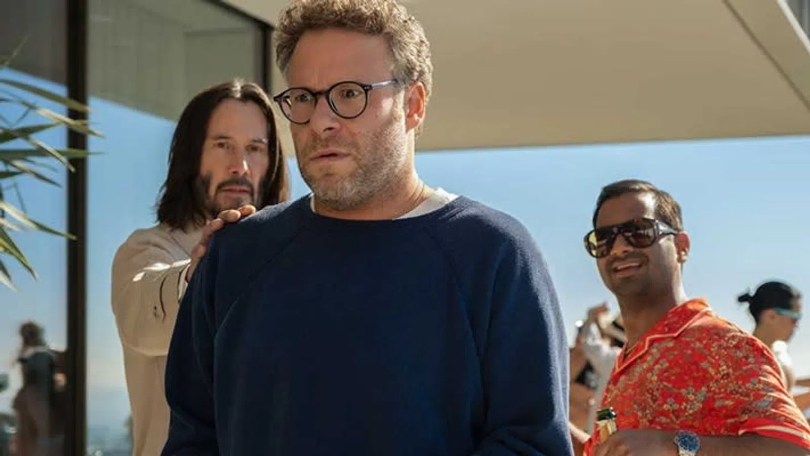 Man in glasses looks worried while others in casual clothes enjoy a sunny outdoor gathering near modern architecture.
