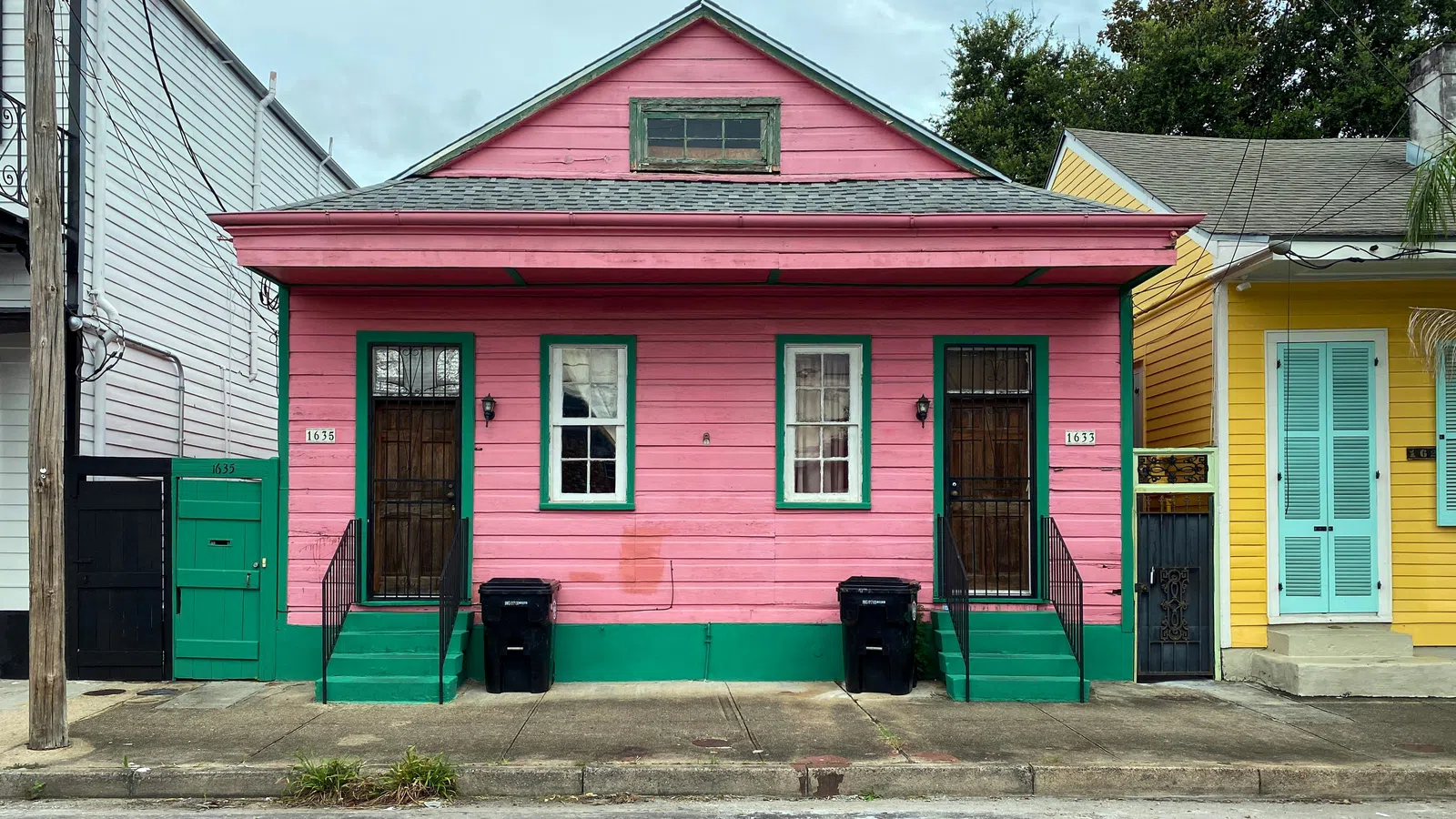 Pink wooden house with green trim, two doors, windows, and trash bins on a city sidewalk in daytime.