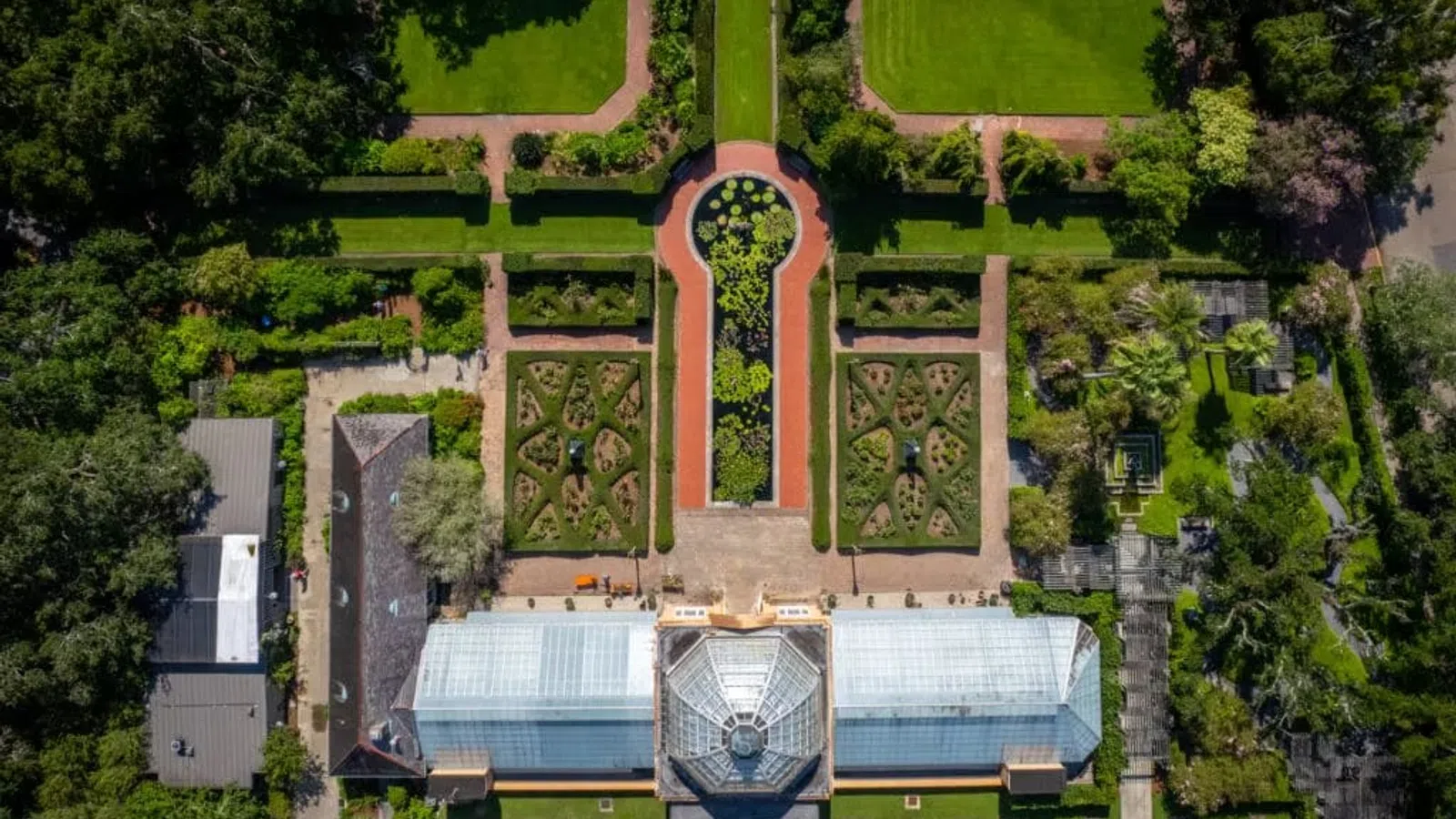 Aerial view of a symmetrical botanical garden with pathways, green lawns, manicured hedges, and a glass conservatory.