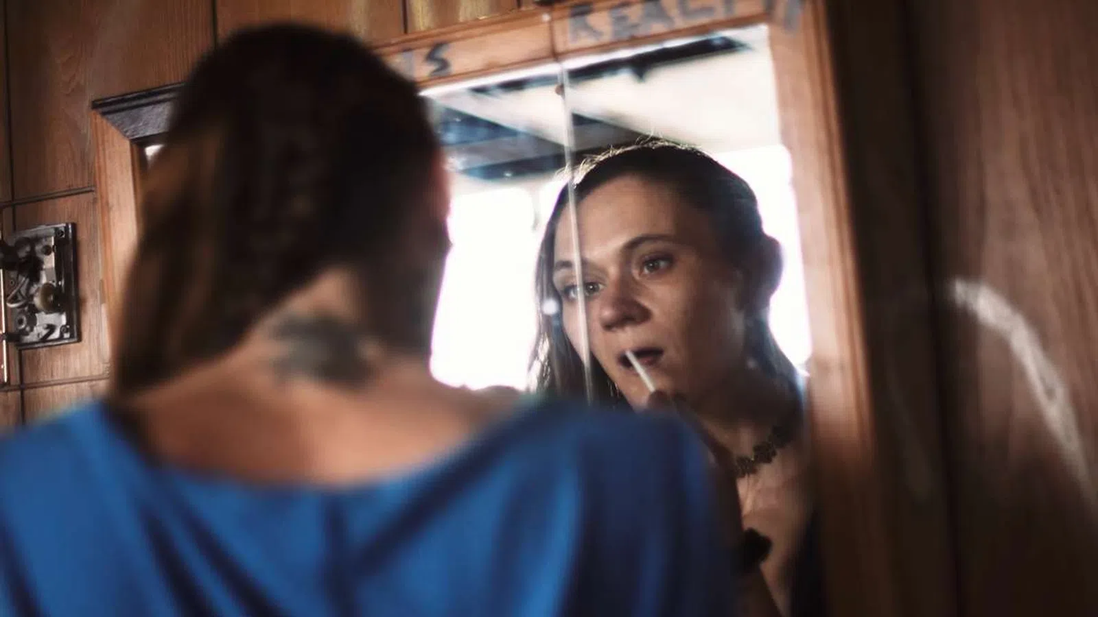 Woman applying lipstick while looking into a vintage wooden mirror in a cozy room.