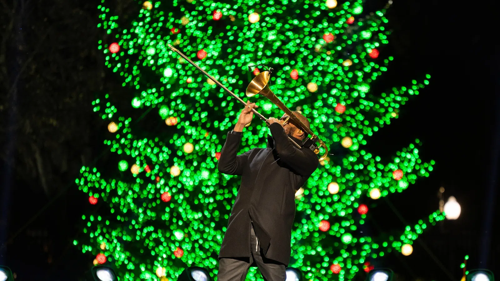 Musician playing trombone in front of a brightly lit Christmas tree with green and red lights at night.
