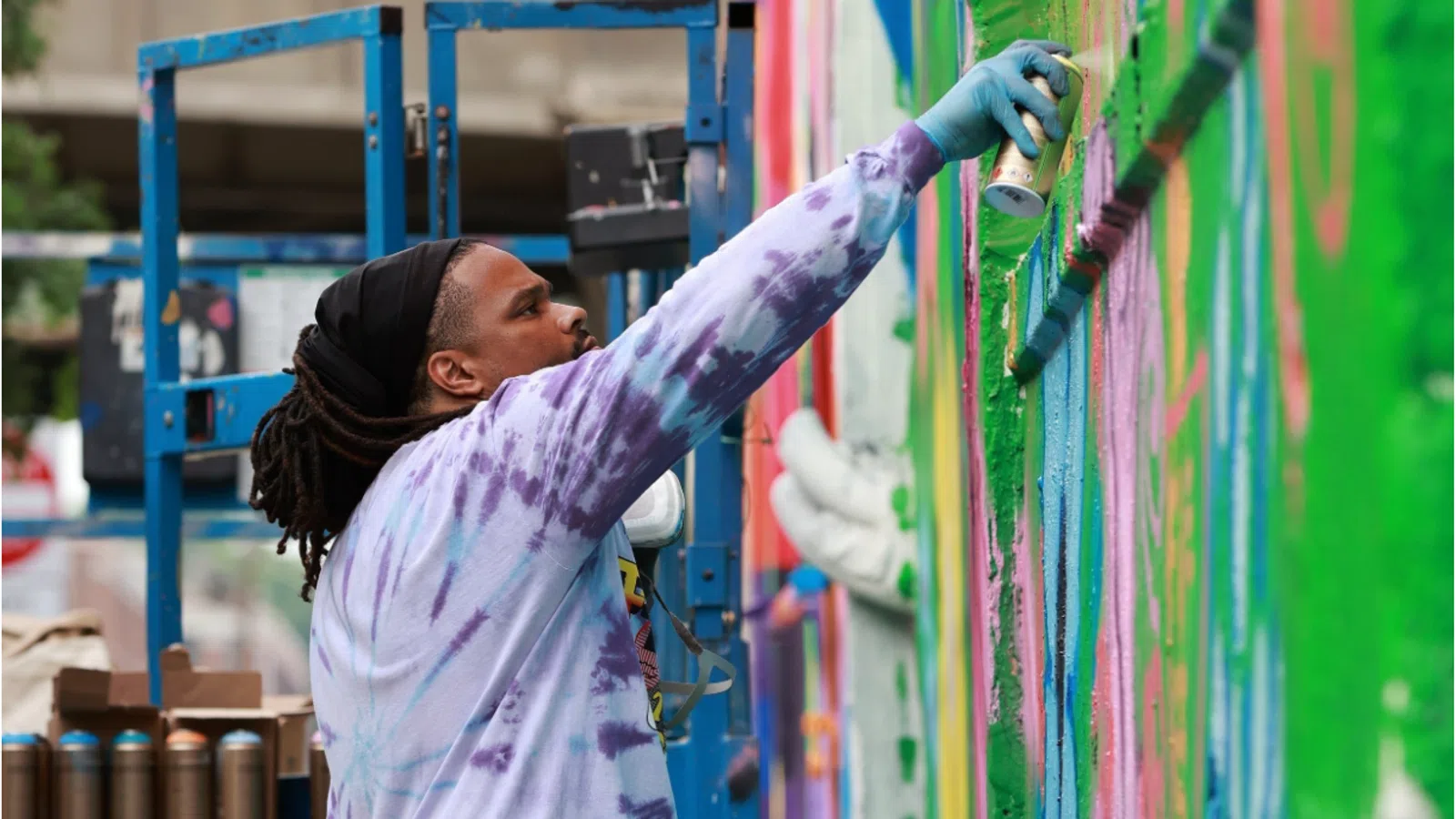 Artist spray painting vibrant graffiti on a large outdoor wall while standing on blue scaffolding in daylight