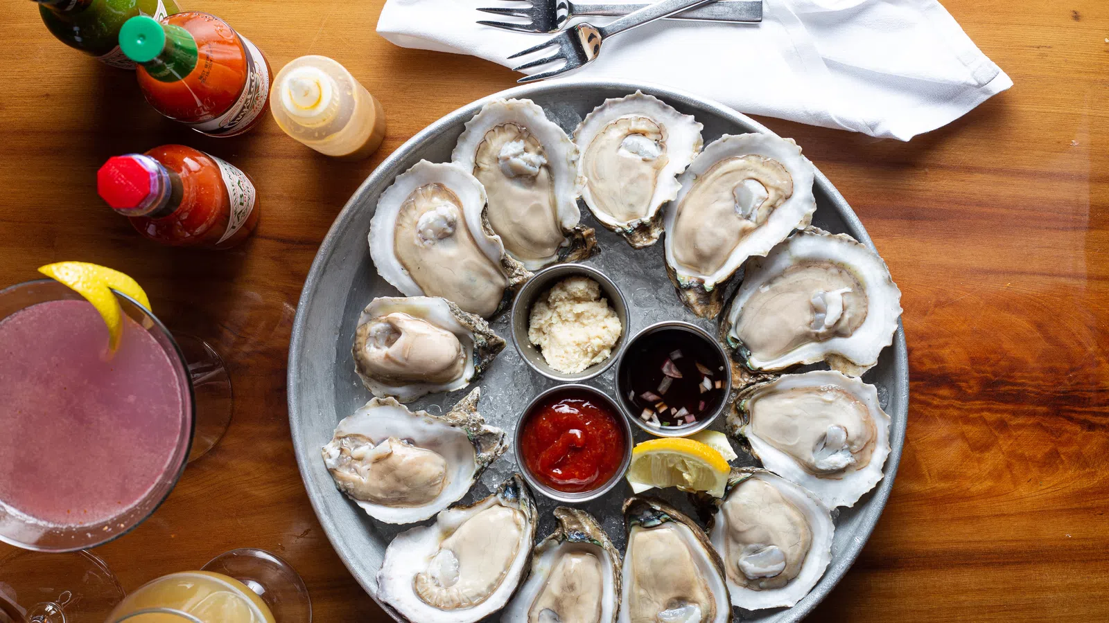 Plate of fresh oysters on the half shell with cocktail sauce, lemon, and condiments on a wooden table with drinks