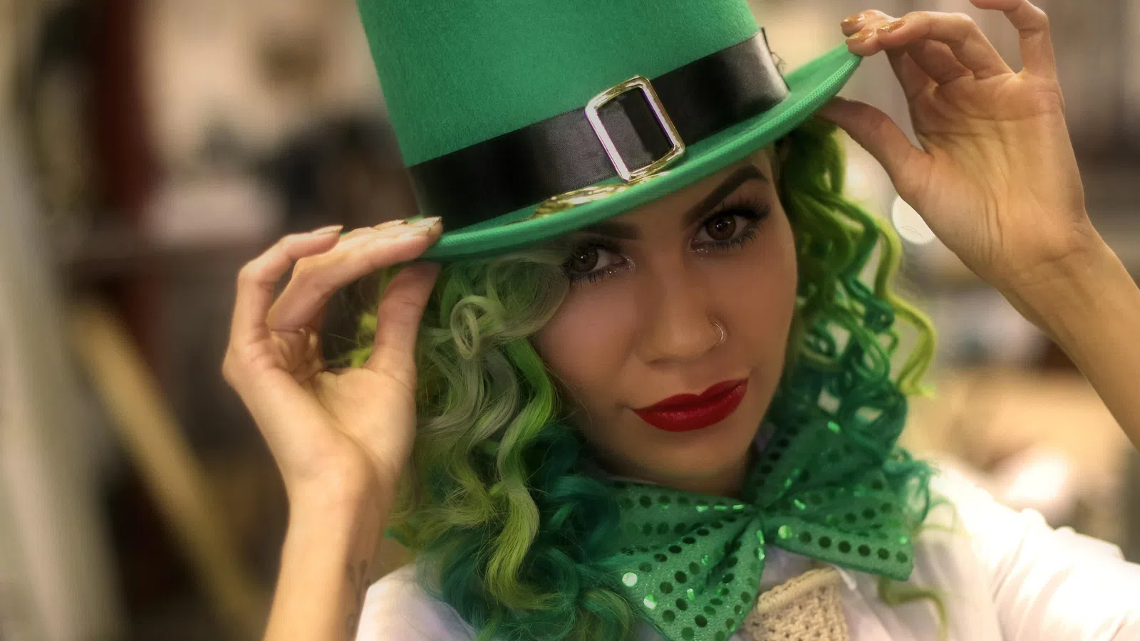Woman with green curly hair wearing a green top hat and matching sequined bow tie, posing indoors.