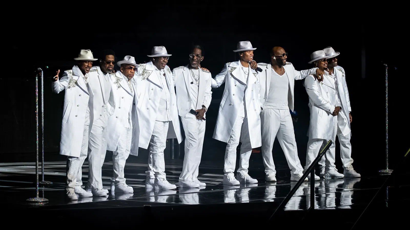 Nine men in coordinated white suits and hats performing together on stage with microphones under spotlight.