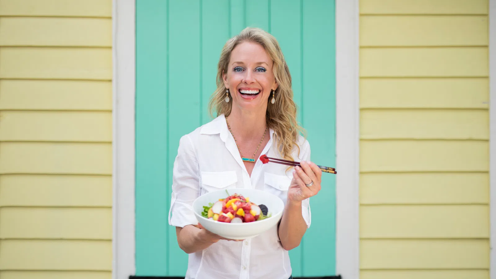 Smiling woman in white shirt holding a bowl of fruit salad with chopsticks in front of pastel yellow and green walls