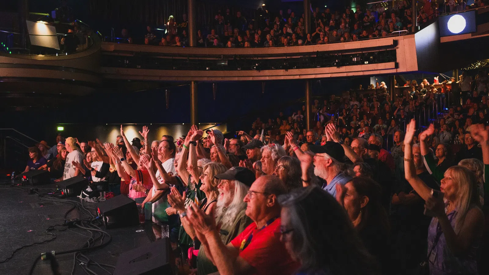 Audience cheering and applauding at an indoor live concert with a multi-level venue filled with spectators