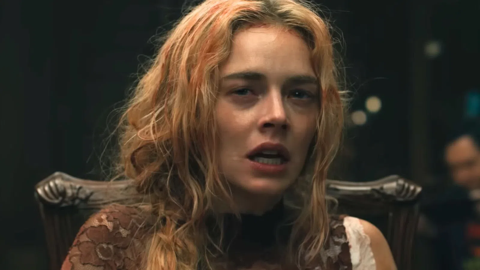 Blonde woman with disheveled hair and distressed expression sitting on an ornate wooden chair in dim lighting.