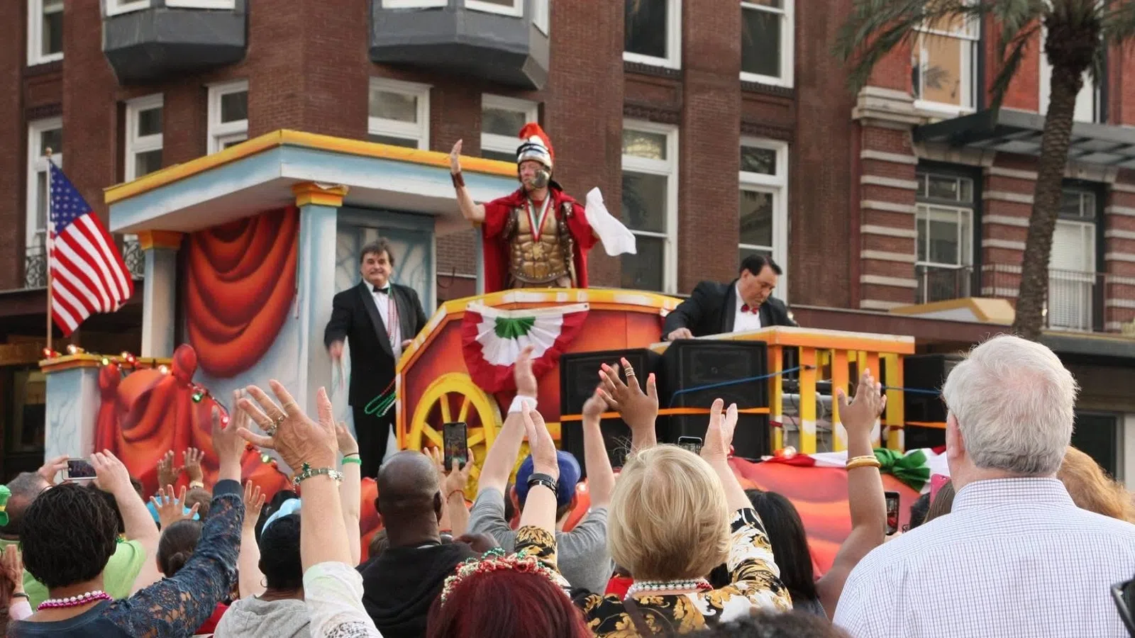 Crowd waves at a parade float featuring a man in Roman armor and two men in suits in a city setting.