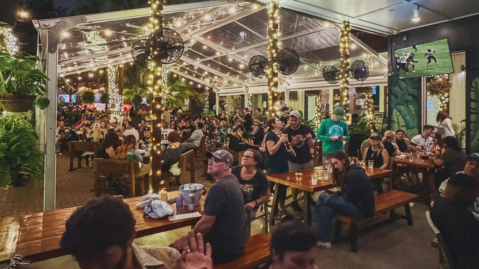 Crowded outdoor bar patio with string lights and people socializing under a transparent roof at night