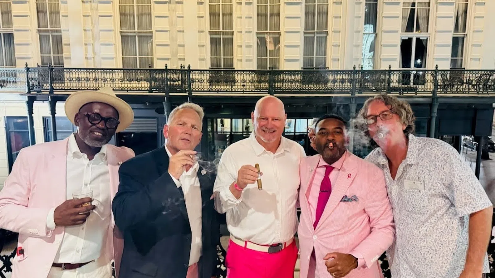 Group of five men dressed smartly enjoying cigars on a balcony with ornate historic building in background