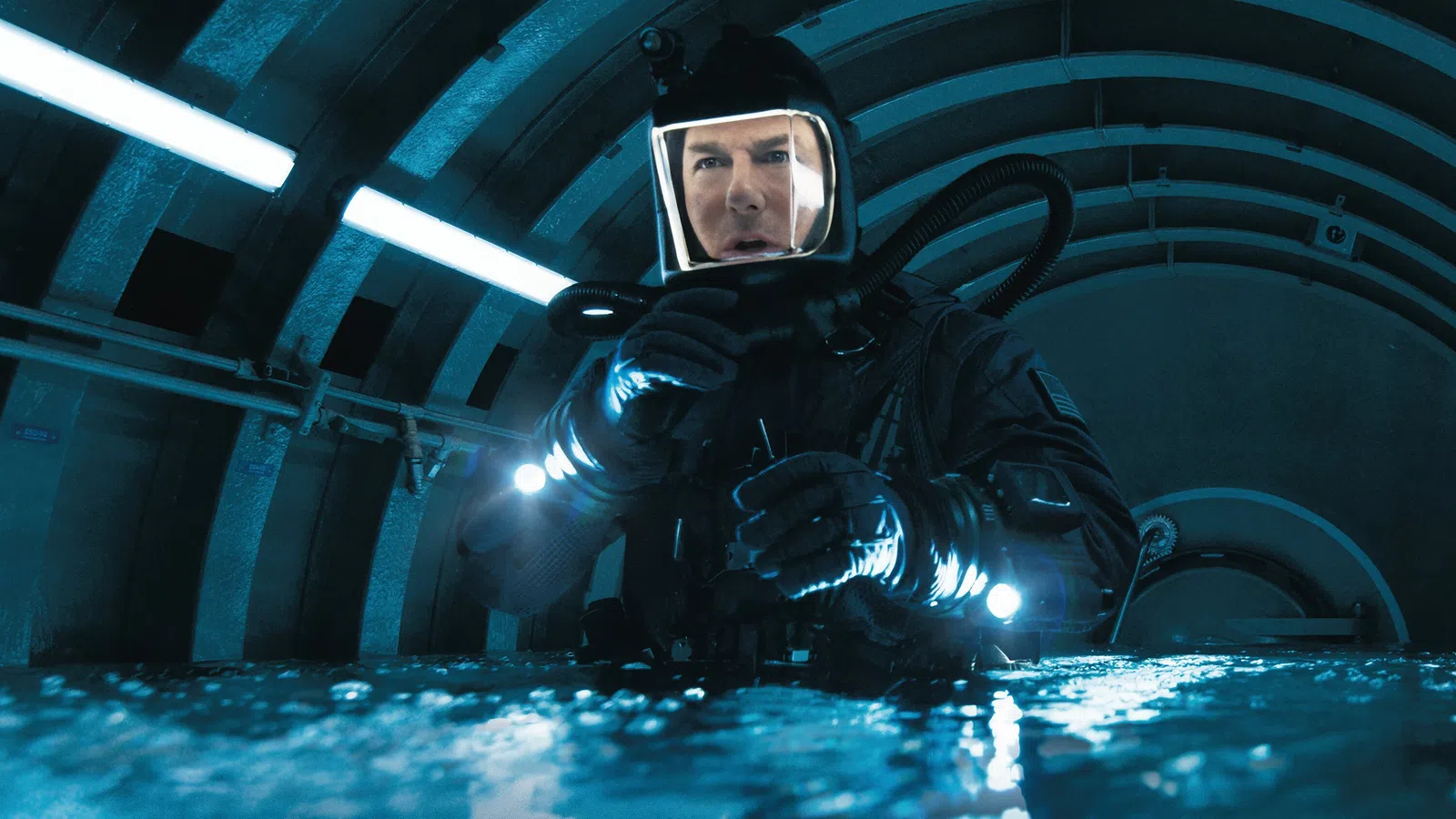 Man in black diving suit and helmet prepares underwater equipment in a dimly lit tunnel.