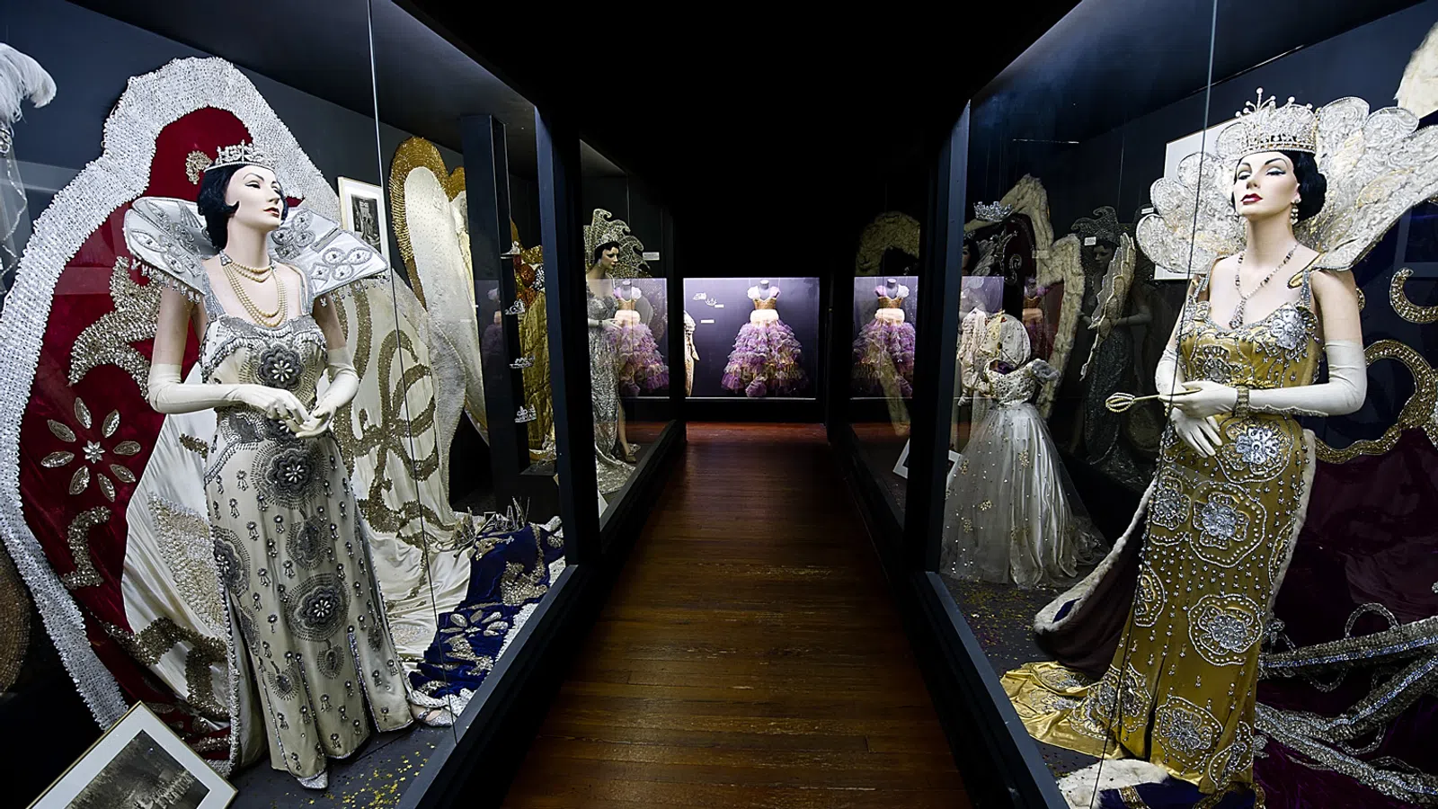 Museum display of elegant vintage mannequins dressed in ornate beaded gowns and royal accessories in glass cases.