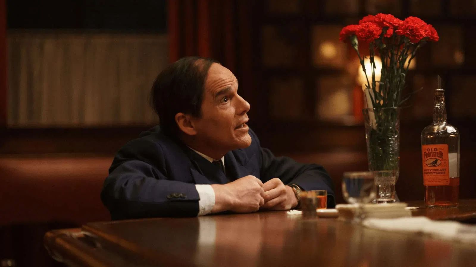 Man in suit sitting at a bar counter with whiskey and red flowers in a dimly lit setting.