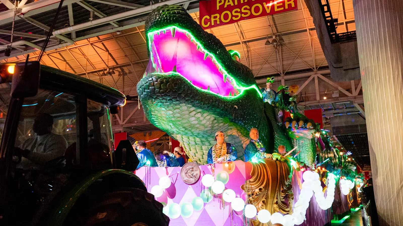 Large illuminated alligator float with purple and green lights at a nighttime parade under a parade crossover sign.