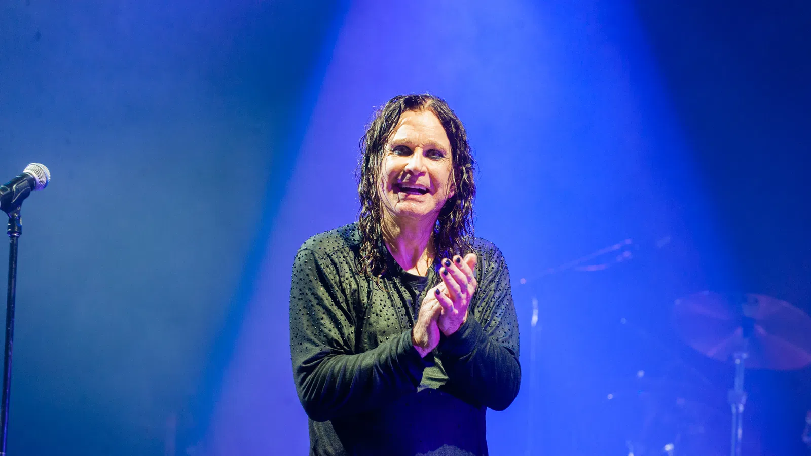 Rock musician with wet hair and black nail polish clapping on stage under blue spotlight with microphone nearby.