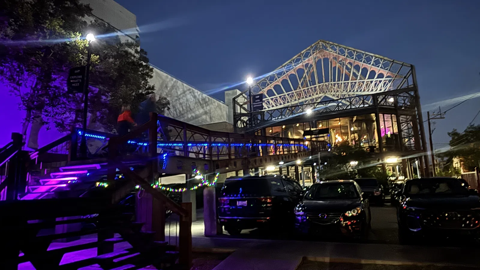 Night view of a modern building with illuminated wooden stairs and parked cars under a steel framework roof structure.